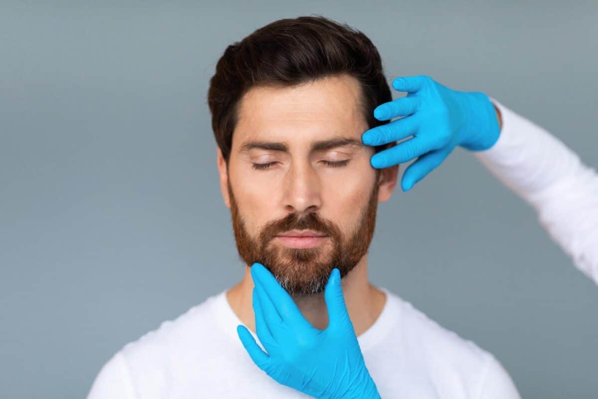 A man with his eyes closed receiving a facial skin assessment for injectable treatment from a doctor wearing blue medical gloves.