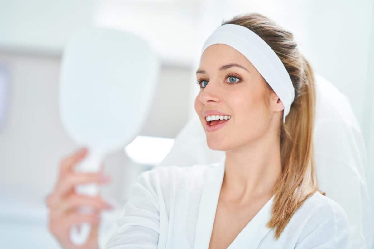 A woman wearing a white headband and dressing gown looks into a handheld mirror, smiling, in a bright, clinical setting after biostimulation treatment for collagen production.