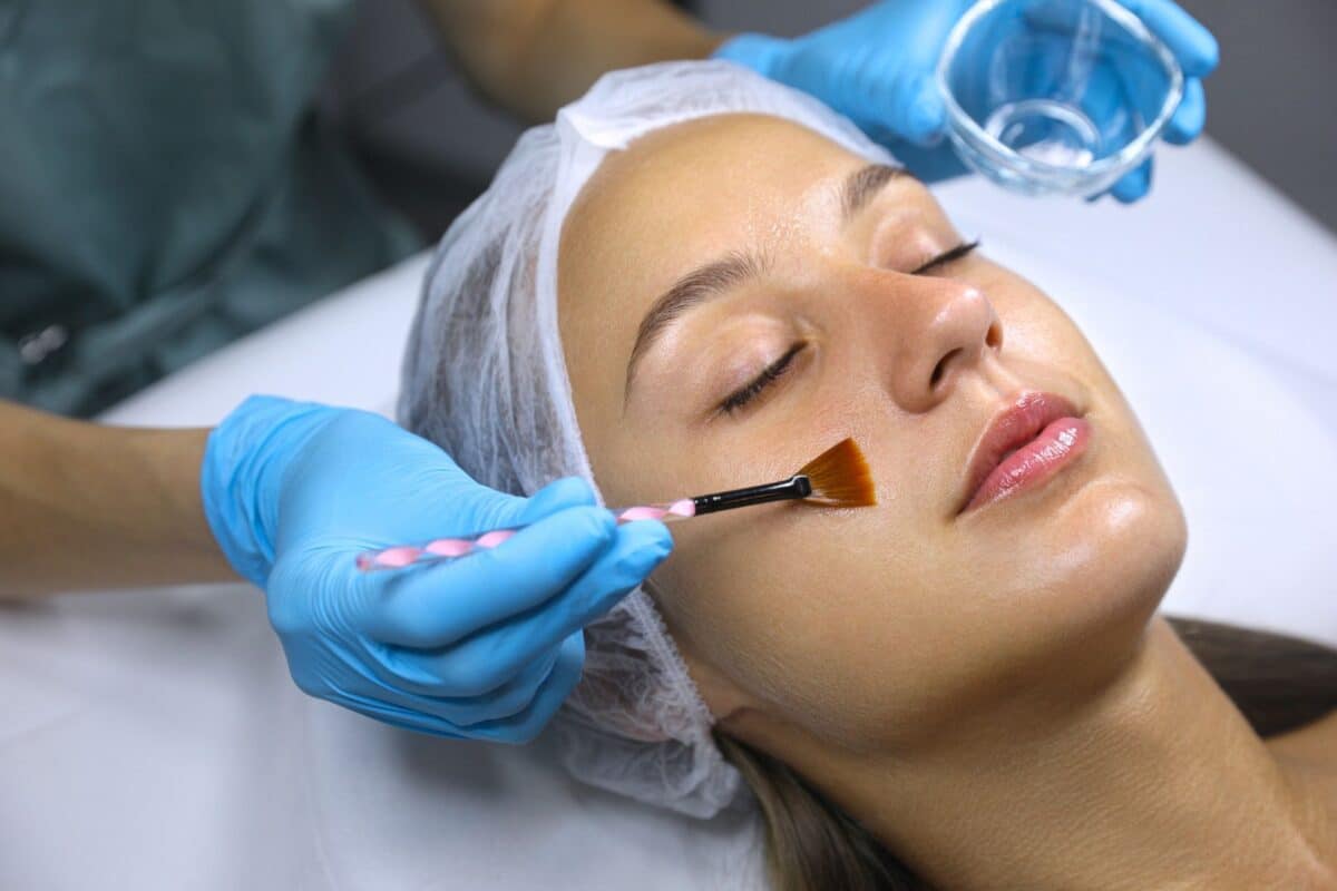 An aesthetician in blue gloves uses a fan brush to apply a chemical peel treatment to a client’s face.