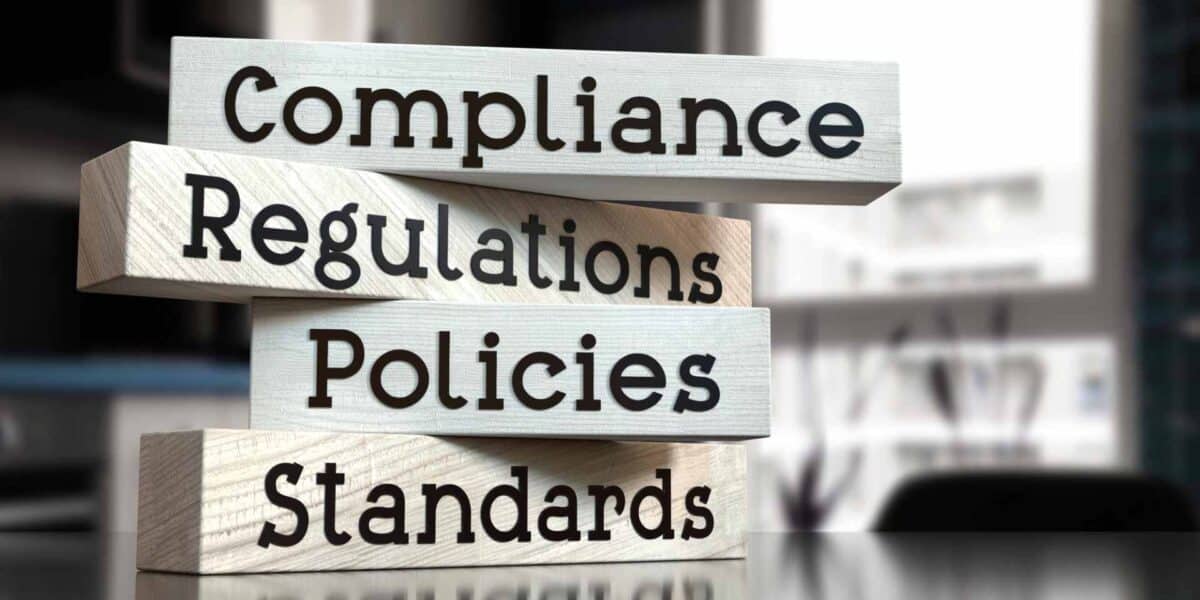 Four wooden blocks stacked vertically display the words: Compliance, Regulations, Policies, and Standards, reflecting compliance-regulatory-in-cosmetic-medicine against a blurred office background.