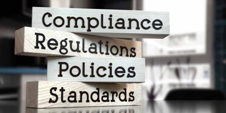 Four wooden blocks stacked vertically display the words: Compliance, Regulations, Policies, and Standards, reflecting compliance-regulatory-in-cosmetic-medicine against a blurred office background.
