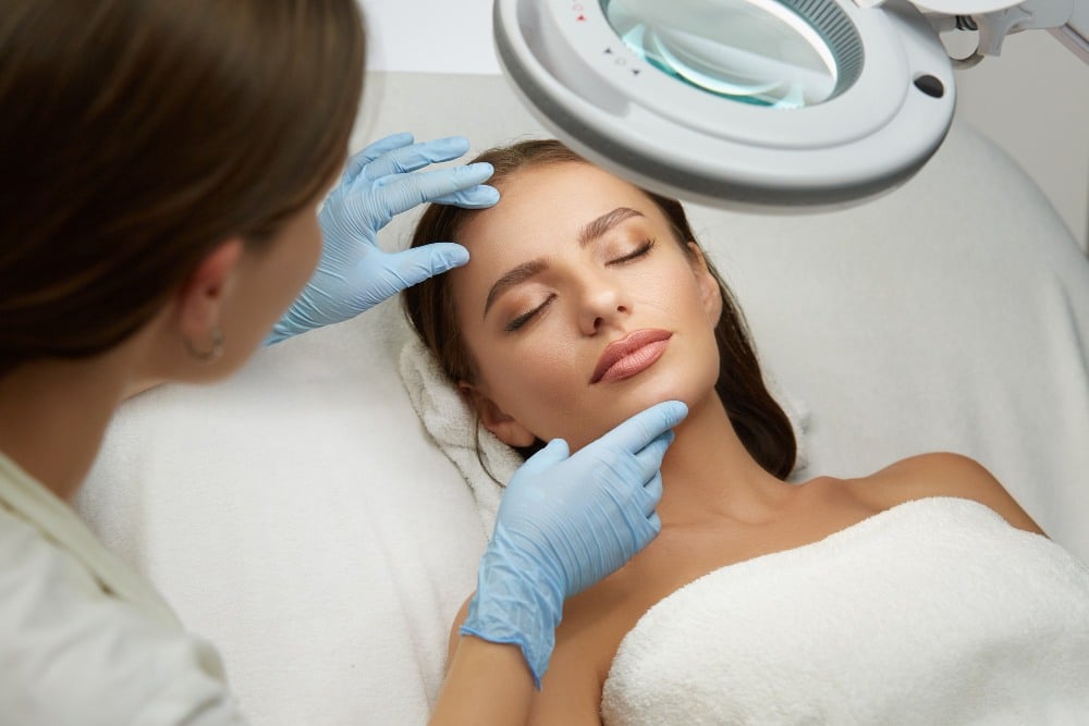 A woman lies on a treatment couch with her eyes closed as a professional wearing blue gloves performs a comprehensive assessment of the face for pan-facial rejuvenation under a magnifying lamp.