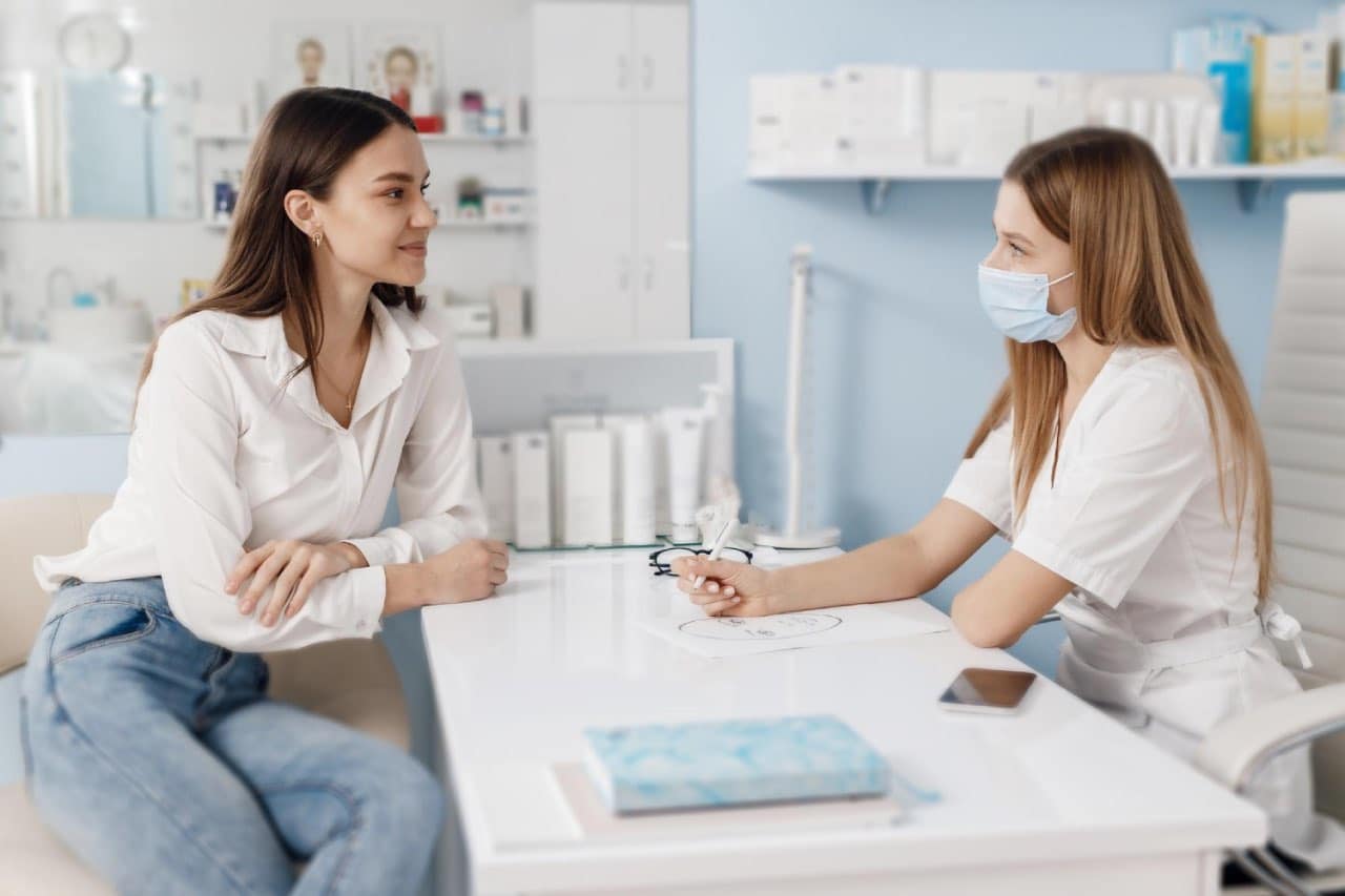 A clinician wearing a face mask consults with a patient at a desk in a cosmetic appointment.
