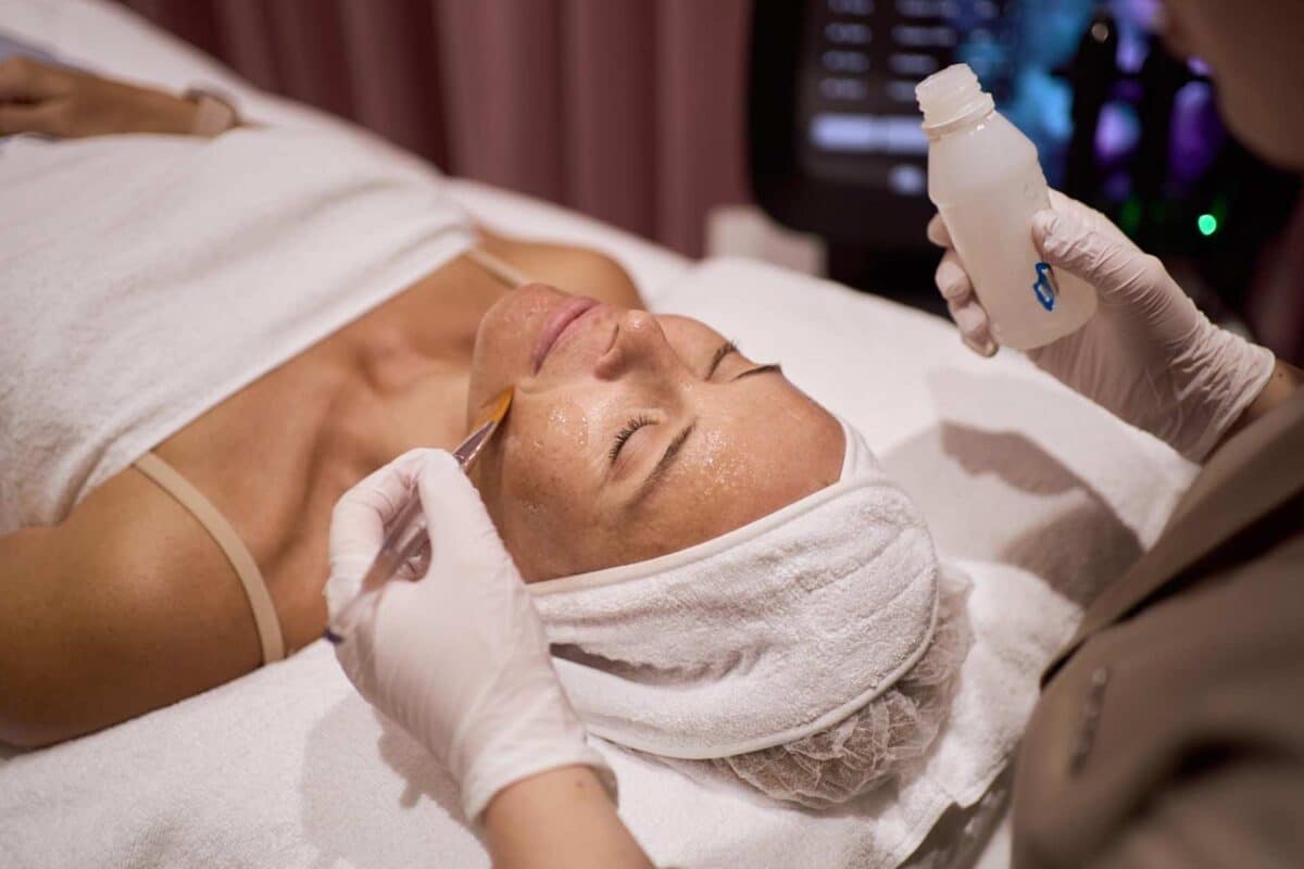 An aesthetician in white gloves uses a brush to apply a cosmetic treatment product to a client’s face.