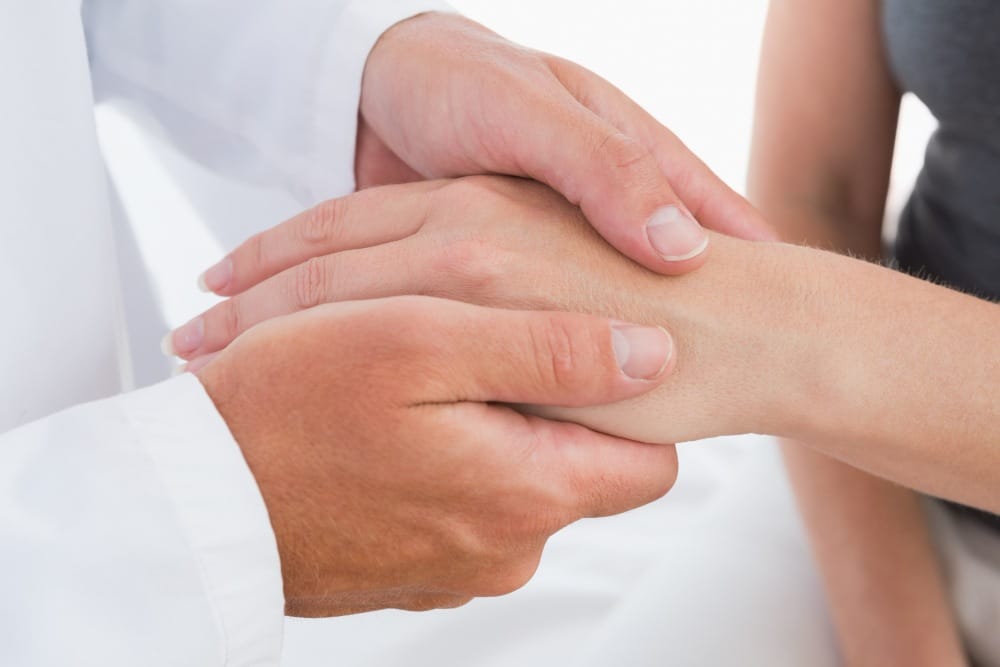 A person in a white coat gently holds another's hand, providing comfort and support as they examine ageing hands for treatments.