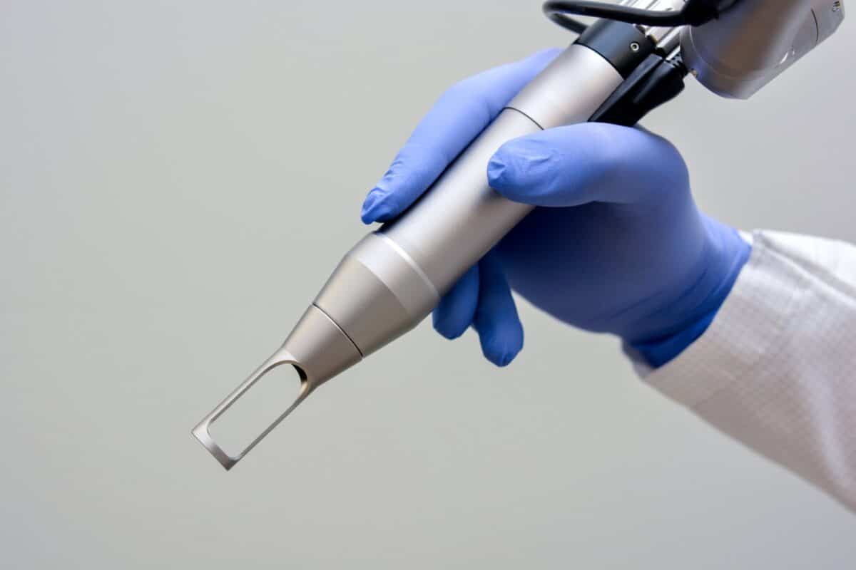 A gloved hand holds a silver medical or cosmetic laser device against a plain background.