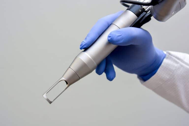 A gloved hand holds a silver medical or cosmetic laser device against a plain background.