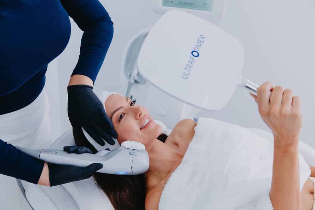 A woman receives a cosmetic treatment on her face with a handheld device whilst holding a mirror.