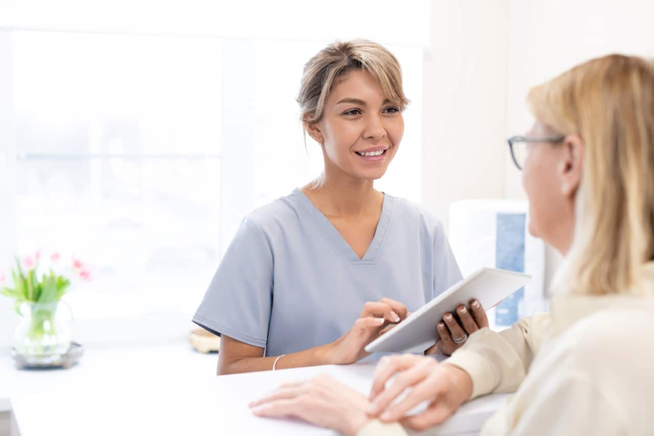 A smiling medical professional in scrubs shows a tablet to a patient during a consultation about injectable risks.