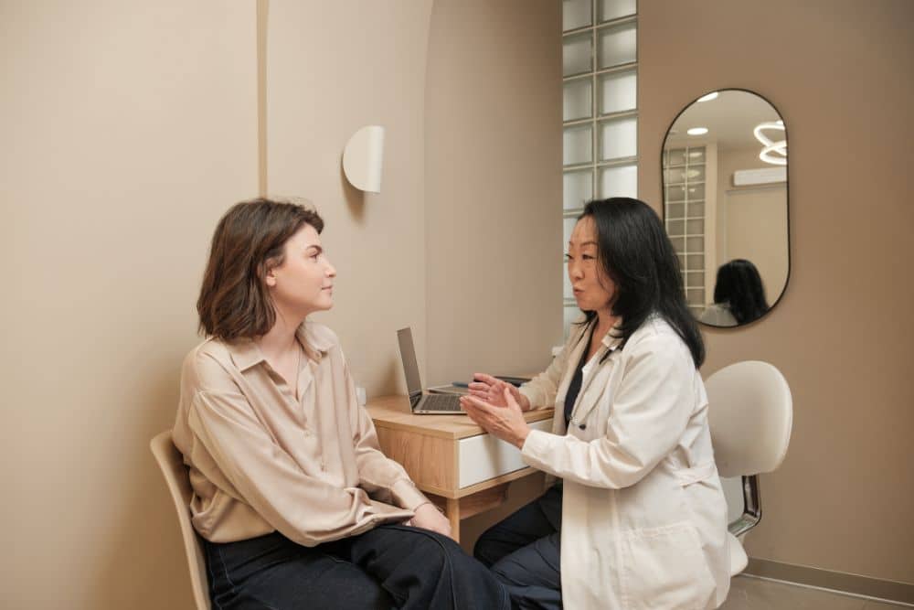 A doctor in a white coat sits opposite a patient during a laser resurfacing consultation with a dermatologist, discussing treatment options in a medical office with neutral-coloured walls and a desk with a laptop.