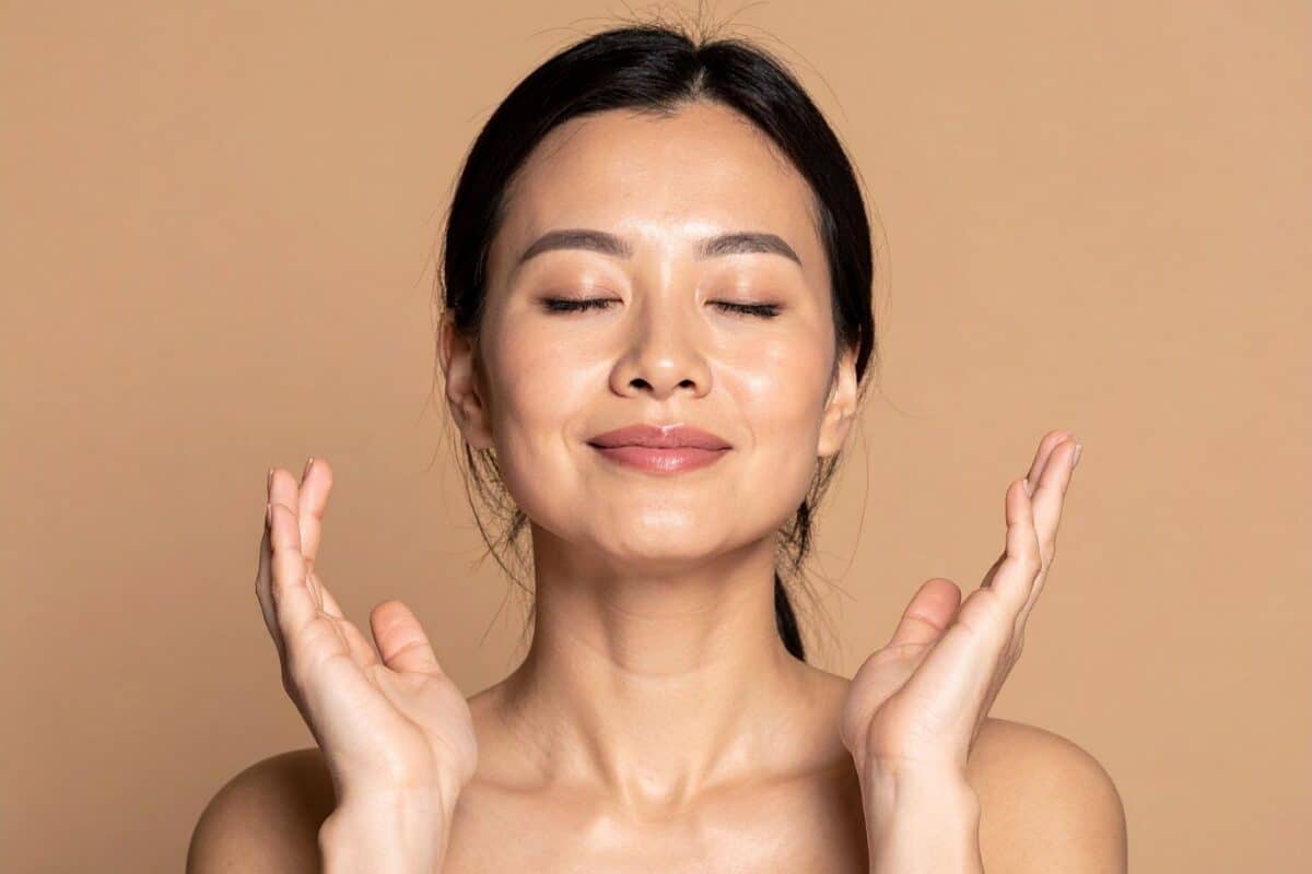 Woman with closed eyes and a slight smile holds her hands near her face, showcasing a calm confidence, possibly reflecting a positive laser resurfacing pigment treatment result against a plain beige background.