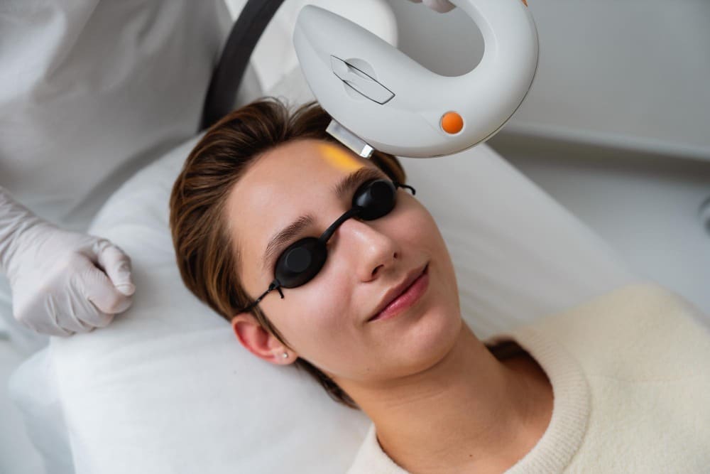 A person wearing protective goggles undergoes a facial treatment with a handheld laser device operated by a gloved technician.