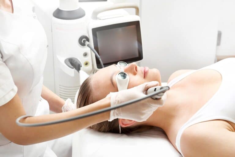 A woman lying on a treatment bed receives a facial procedure with a handheld device from a gloved professional in a clinical setting.