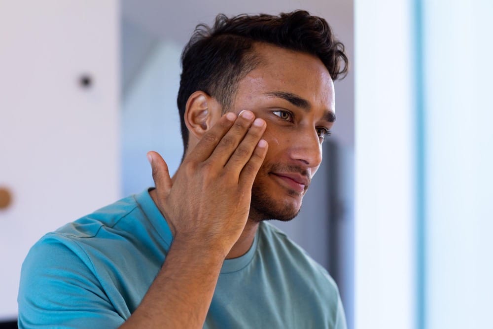 A man in a teal shirt touches his cheek with his hand, looking at his reflection in a brightly lit room.