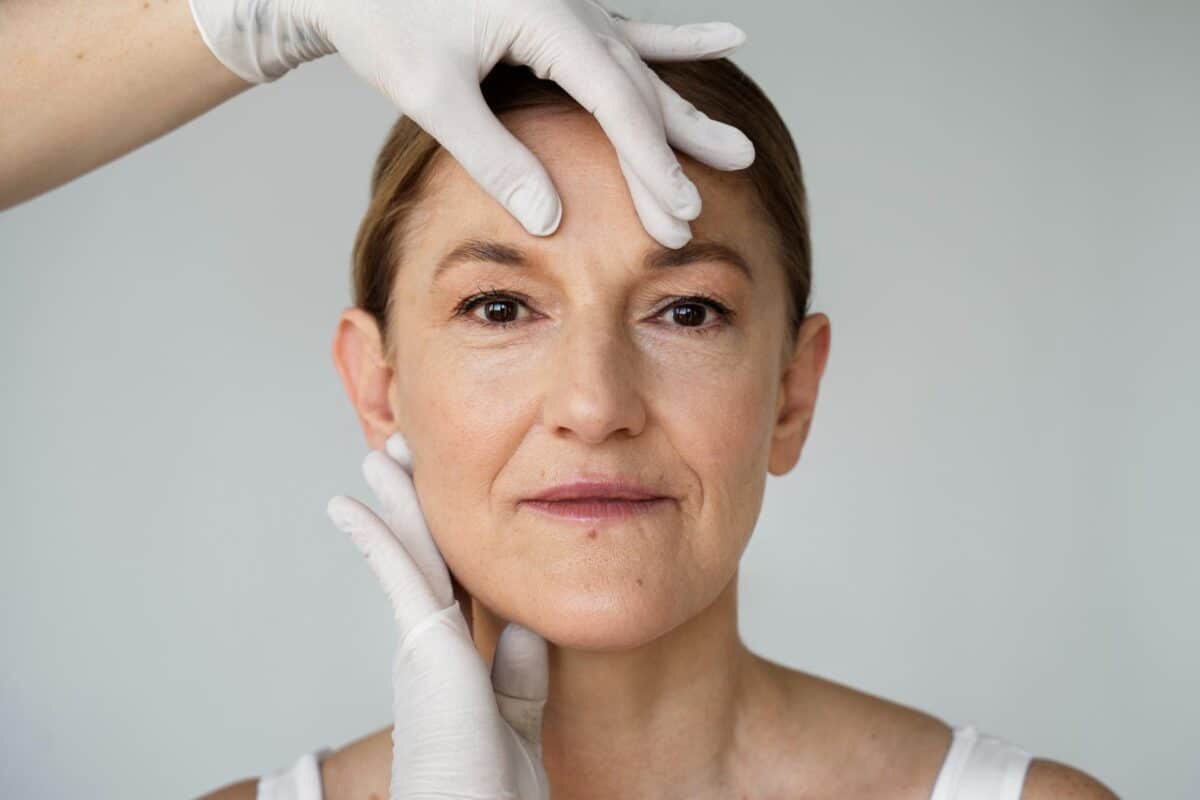 A woman with a neutral expression has her face gently examined by gloved hands, possibly during a cosmetic or dermatological consultation for a Non-Ablative Fraxel Laser treatment.