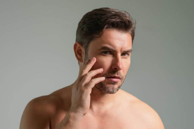 Shirtless man with short brown hair and light stubble touches his cheek, looking thoughtfully at the camera against a plain background—considering the benefits of Fractional Light-Based Treatments for smoother skin.