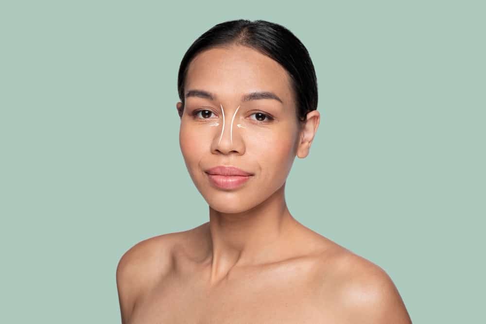 Woman with bare shoulders facing forward, neutral expression, with white contour and highlight make-up lines suggesting non-surgical rhinoplasty on her nose and under-eye area, set against a plain light green background.
