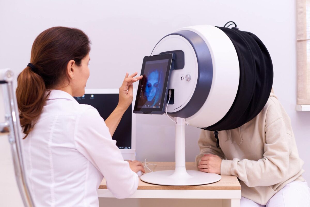 A clinician uses a tablet to analyse a facial scan for pan-facial-rejuvenation-comprehensive-assessment-of-the-face, while a patient sits with their head in a circular medical imaging device.