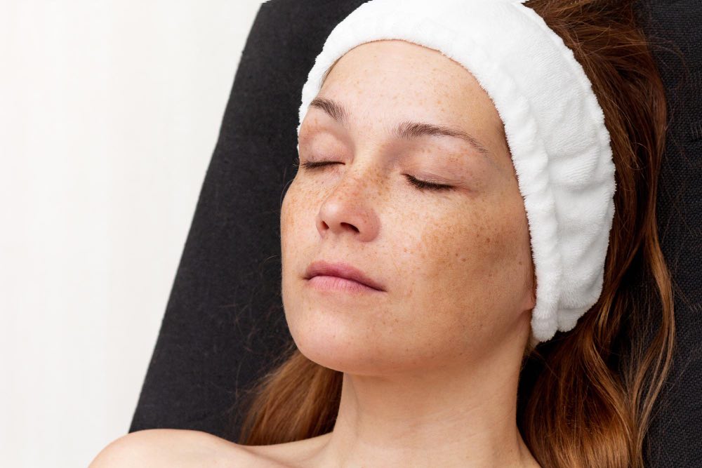 A woman with freckles and a white hairband reclines with her eyes closed, appearing relaxed, against a dark background.