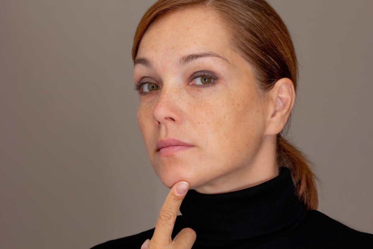 A woman with light skin and freckles touches her chin whilst looking directly at the camera. She is wearing a black polo neck and has her hair tied back.