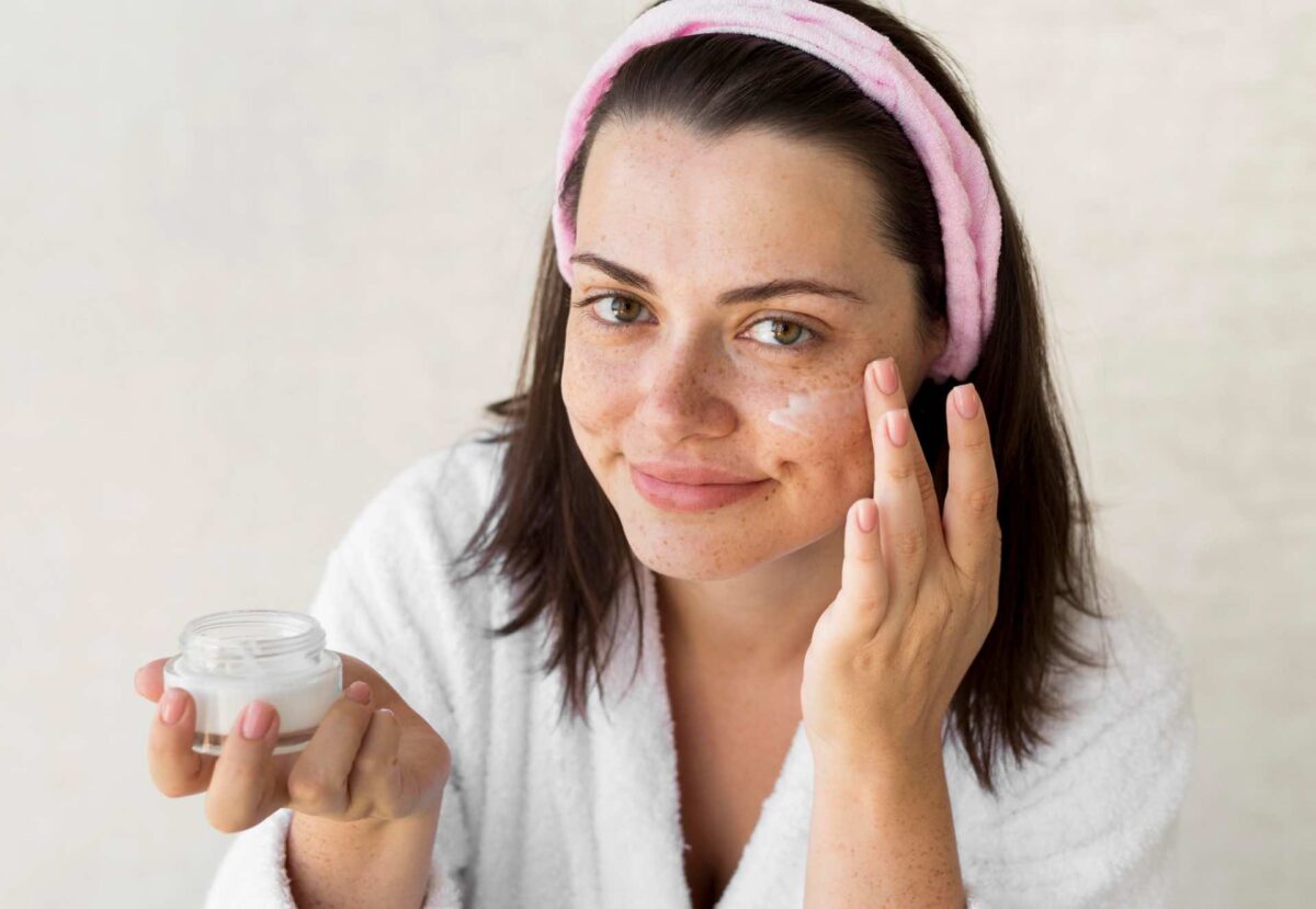 Woman in a white dressing gown and pink headband applies cream to her face—an essential step in pre-laser-resurfacing-treatment-skin-conditioning—holding a small jar of skincare product and smiling at the camera.