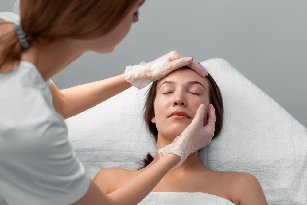 A woman wearing gloves examines the face of a woman lying on a treatment table with her eyes closed, highlighting the role-of-anaesthetic-in-patient-comfort during cosmetic treatments.