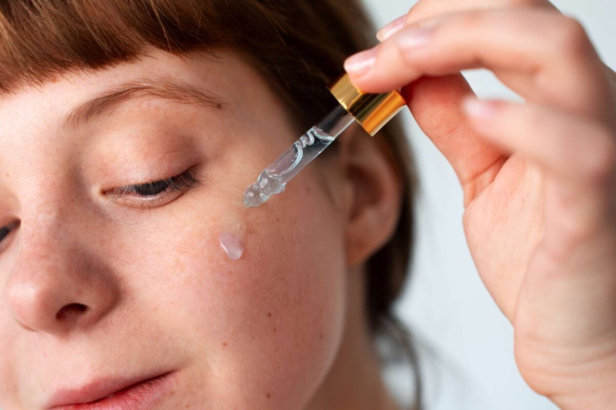 A hand uses a glass dropper to apply a clear peptide facial serum to a person’s cheek.