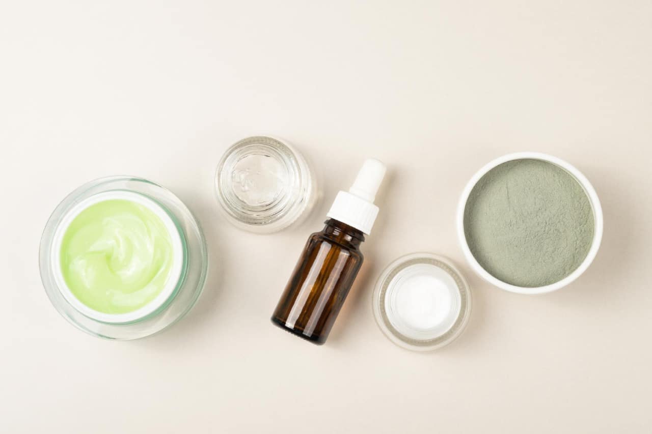 Top-down view of skincare products, including a jar of green peptide cream, an amber dropper bottle of peptide serum, and a bowl of green clay powder.