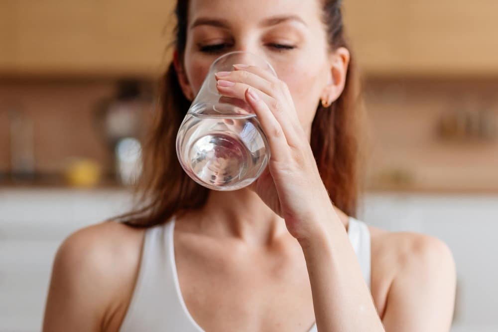 A woman in a white top drinks a glass of water to stay hydrated for first cosmetic treatment. 
