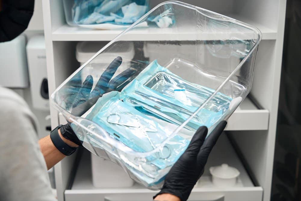 Person wearing black gloves holding a clear container filled with individually wrapped sterilised equipment before aesthetic procedures in a clinical setting.