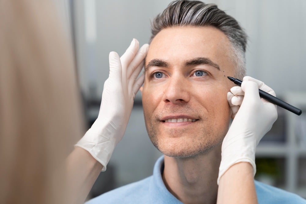 A man with grey hair is having marks drawn on his face with a black marker by a person wearing white gloves, possibly as part of a thorough facial assessment before an aesthetic procedure.