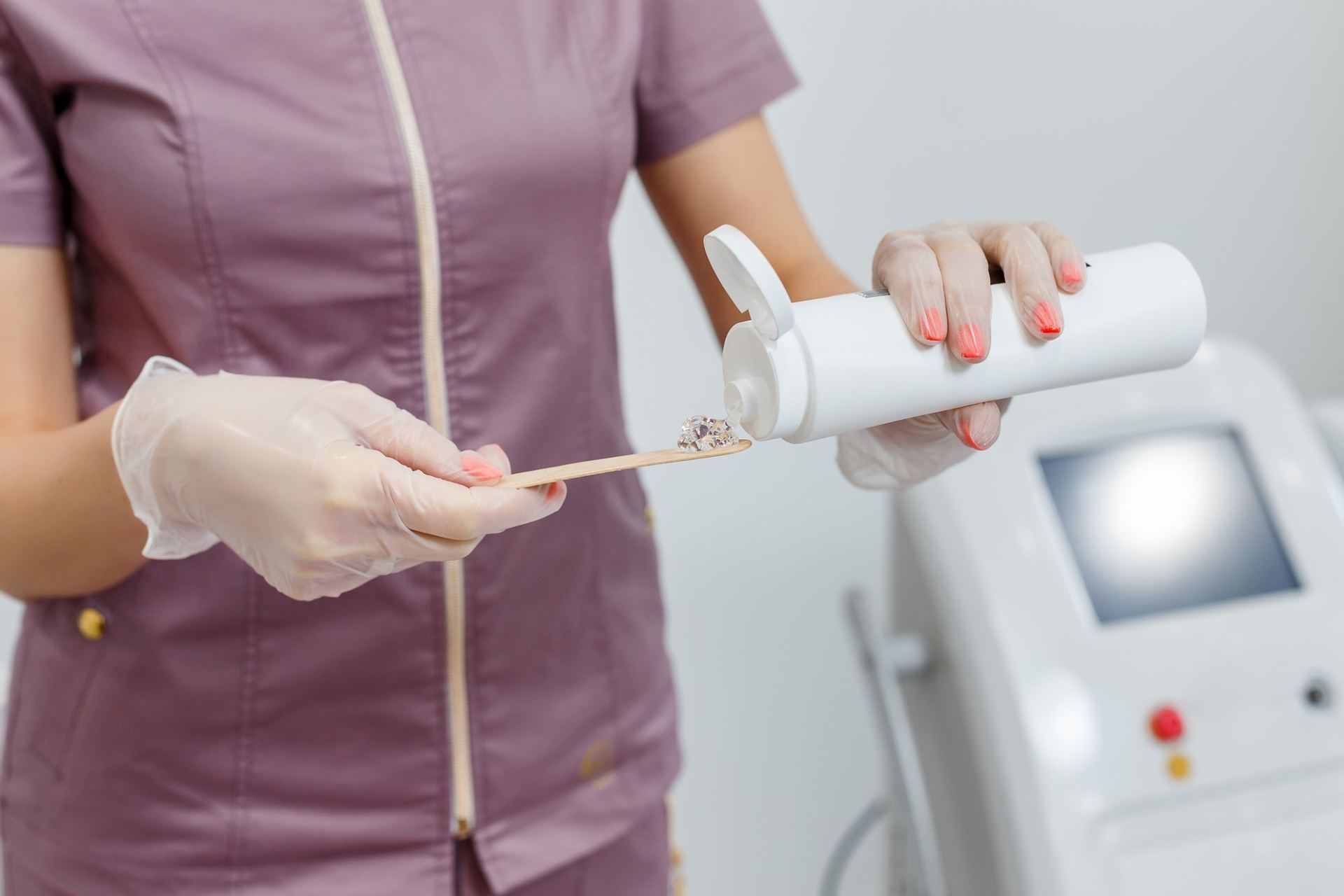 A nurse wearing gloves dispenses topical anaesthetic cream onto a wooden spatula from a white bottle, with medical equipment visible in the background before a cosmetic treatment.