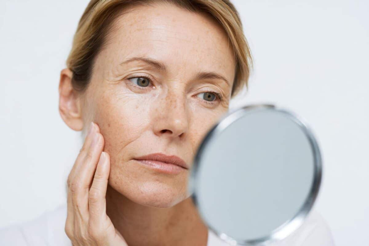 A woman looks into a hand-held mirror while touching her cheek, appearing to examine her skin closely for signs of melasma.