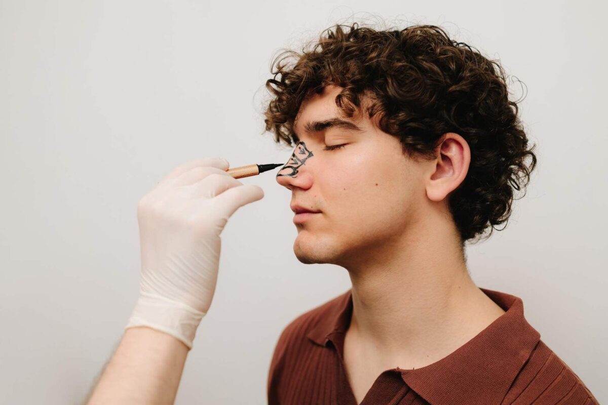 A person with closed eyes has a drawing made with black eyeliner on their nose by a gloved hand, illustrating the planning process for a Non-Surgical Rhinoplasty.