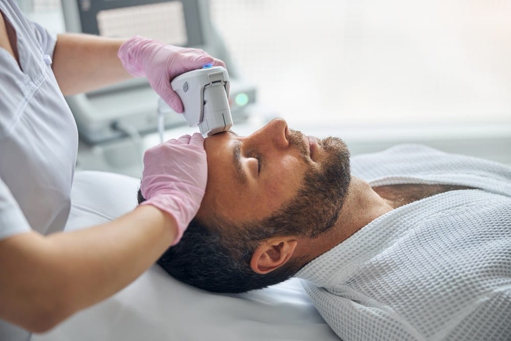 A person in a white dressing gown receives a facial skin treatment with a handheld device from a professional wearing pink gloves.