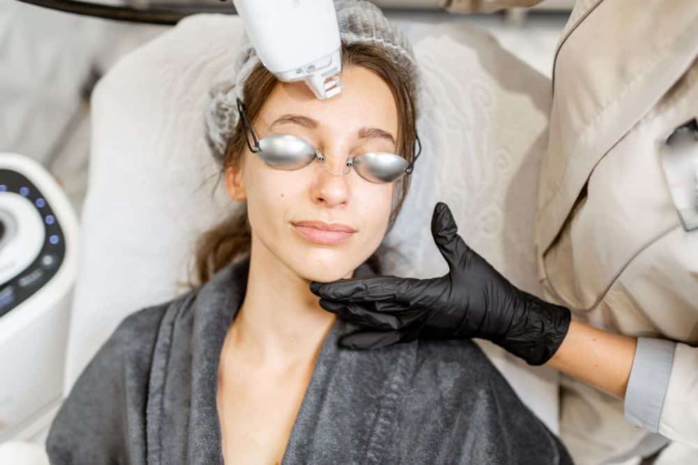 A young woman undergoes laser resurfacing, wearing protective goggles as a gloved professional delivers a facial treatment with a handheld device.
