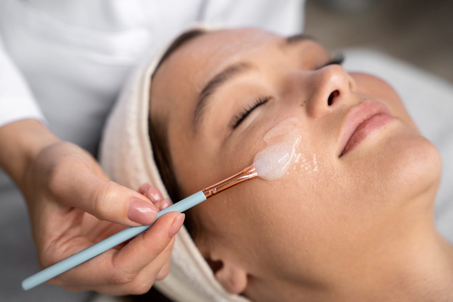 A person with closed eyes receives a facial treatment as a clear gel is applied to their cheek with a brush by a beautician.