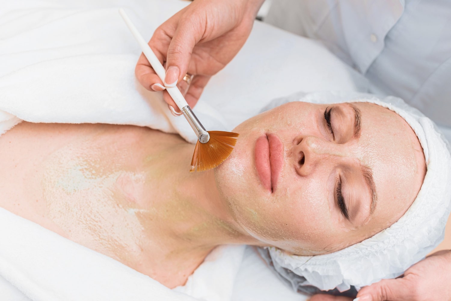 A person lying down with eyes closed receives a facial treatment, as a brush is used to apply a masque to their face and neck.