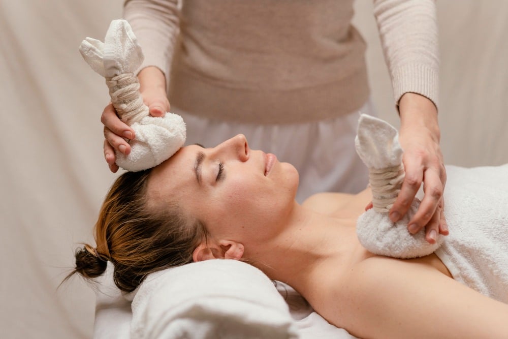 A person lies on a massage table with eyes closed while another applies herbal compresses and ice bags to improve comfort of patient during an aesthetic procedure, focusing on the forehead and shoulder.