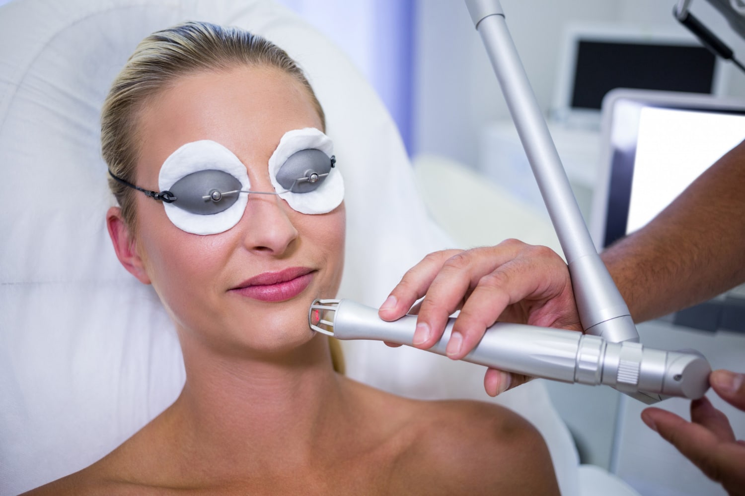 A woman with eye protection undergoes a cosmetic laser treatment on her face in a clinical setting.