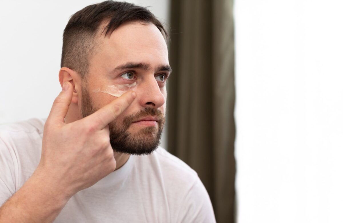 A man with a beard applies cream under his right eye using his finger as part of laser aftercare and healing of skin.