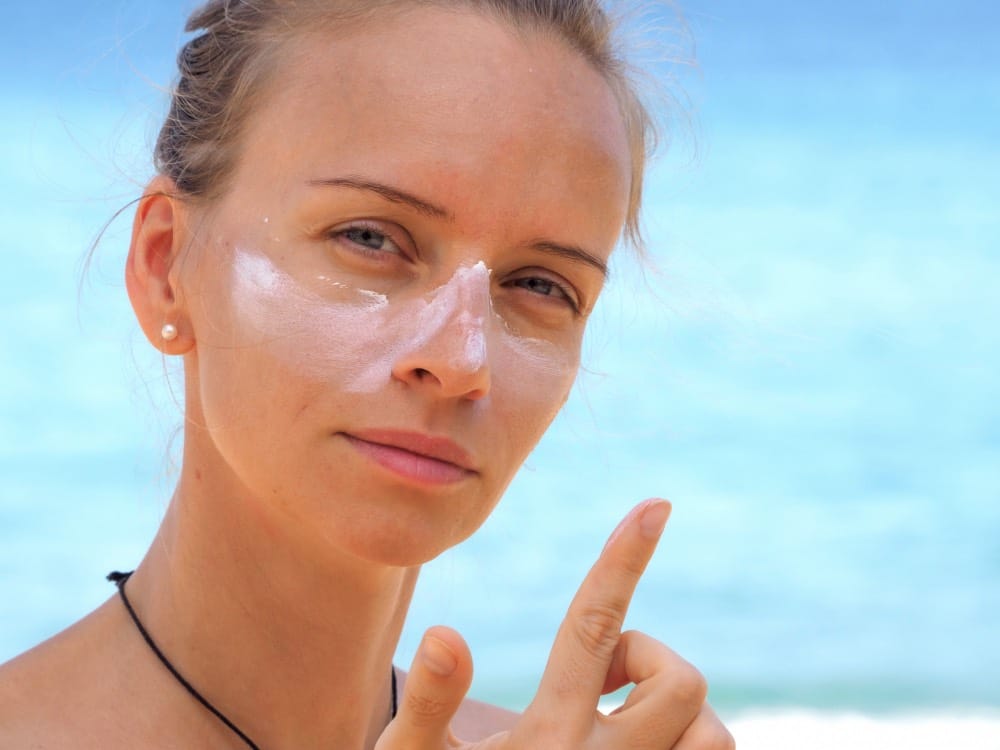 A woman at the beach applies suncream to her face, with visible suncream on her nose and cheeks, highlighting the importance of sun protection and medical-preparations-sun-protection-before-laser-resurfacing. She points her finger upward.