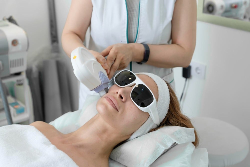 A woman wearing protective goggles receives a pigment-treatment-IPL laser skin procedure on her face from a technician in a clinical setting.